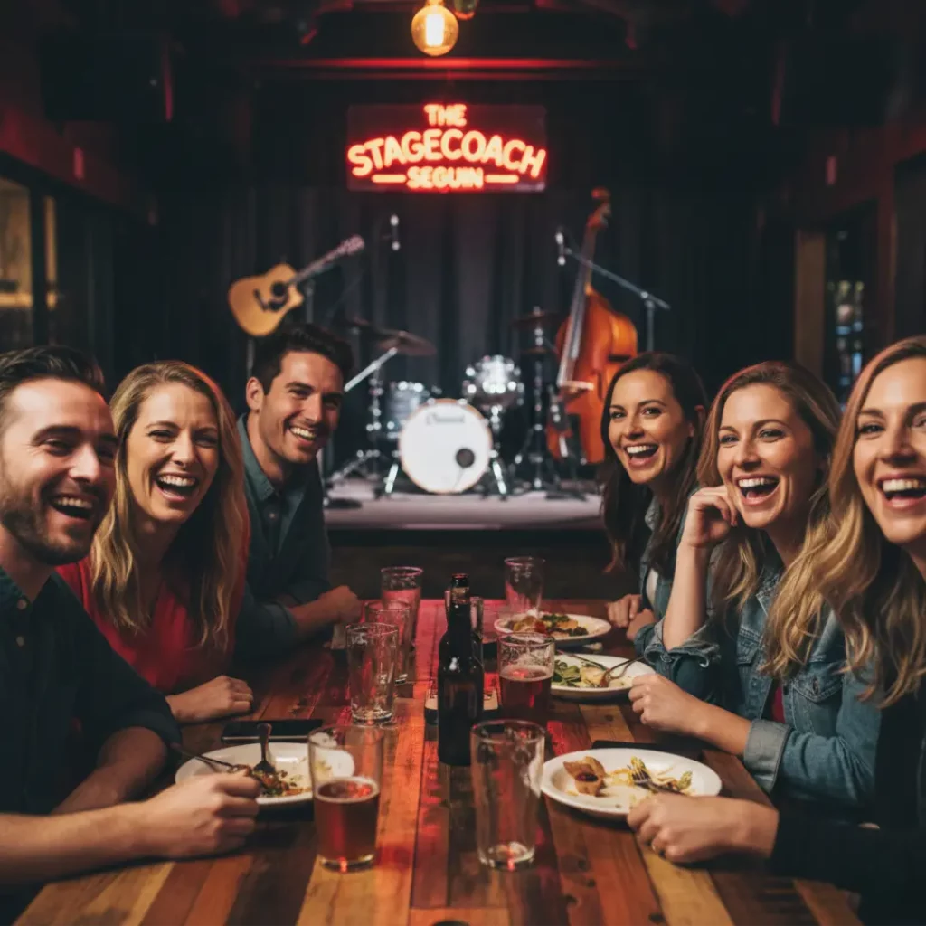 A group of friends smiling at a long wooden table with beers and food, positioned in front of a small indoor stage with instruments and a neon sign.