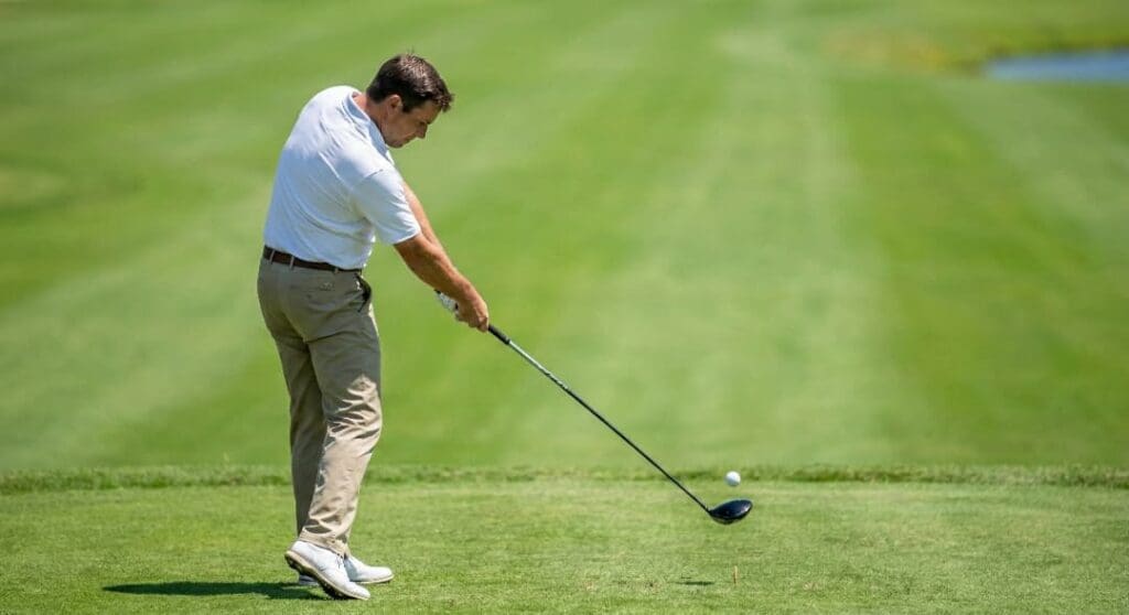 A man in a white polo shirt and tan pants follows through on a golf swing at a tee box.